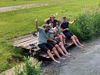 Fünf Personen sitzen auf einem Holzsteg am Wasser, haben die Füße im Wasser und zeigen Daumen nach oben